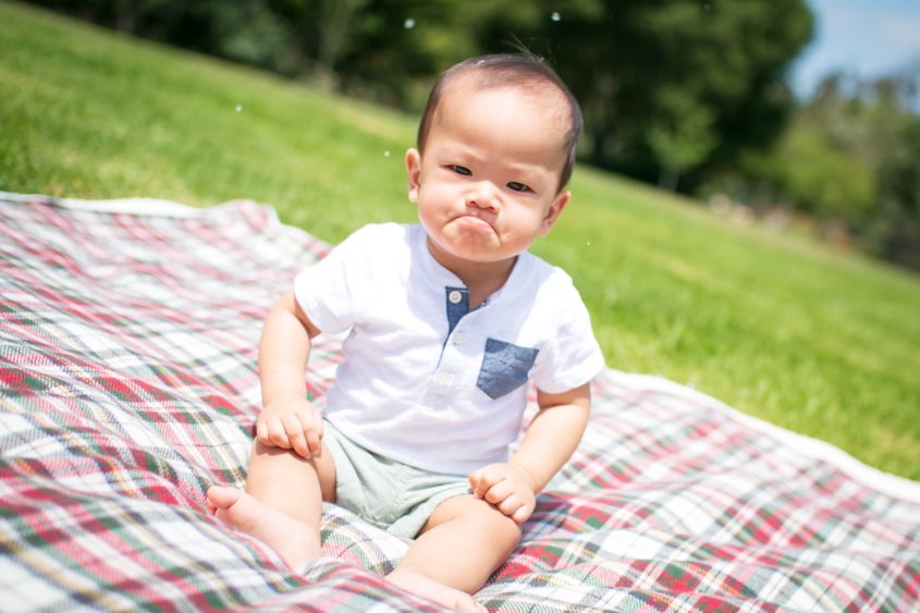 A baby pouts on a blanket in a park.